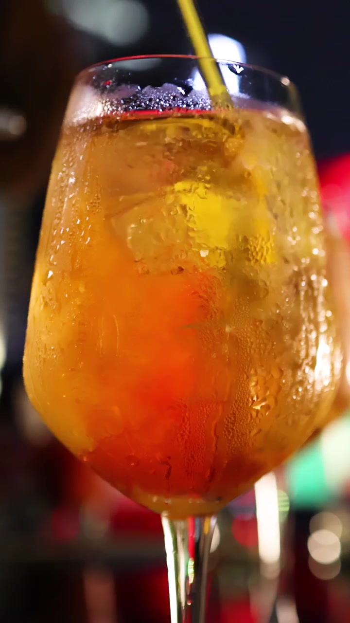 Primo piano di un cocktail all'arancia ghiacciato in un bicchiere a calice, mescolato con un bastoncino giallo sotto una vivace illuminazione da bar, che evidenzia la condensa e la guarnizione di agrumi.