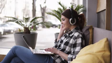 Beautiful,-cheerful-woman-listening-to-music-on-laptop-at-trendy-room-with-headphones-sitting-on-a-couch-in-casual.-Happy,-enjoy-her-time.-Side-view