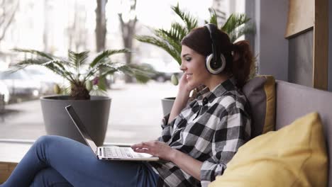 beautiful, cheerful woman listening to music on laptop at trendy room with headphones sitting on a couch in casual. happy, enjoy her time. side view