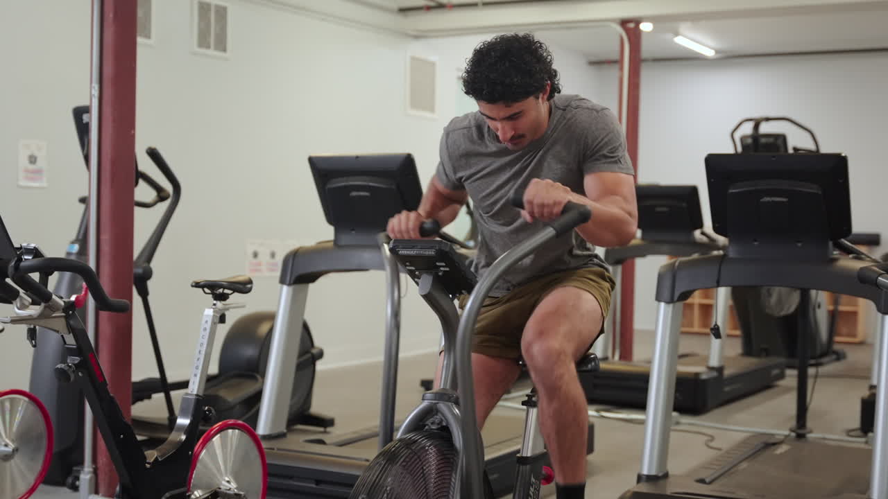 Fixed shot of a man performing an intense air bike sprint in a gym, focusing with determination and looking toward the camera