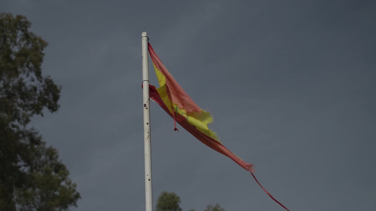 Vecchia bandiera spagnola sbiadita e stracciata, sfilacciata su un'asta bianca contro un cielo grigio coperto.