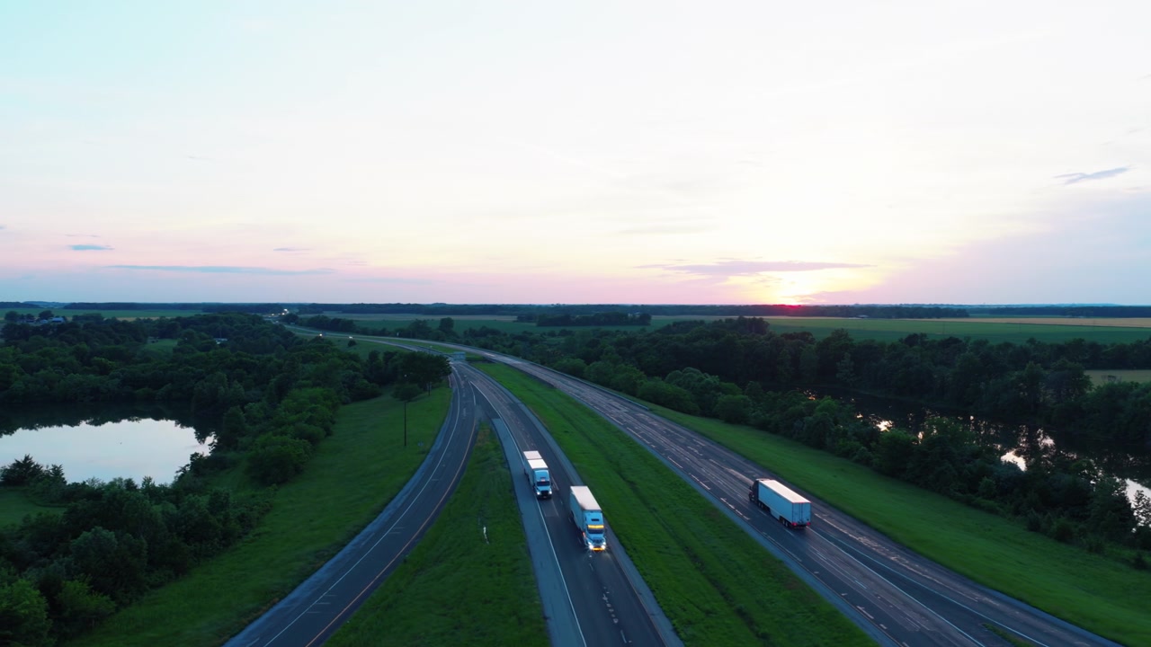 Ilmakuva rahtia kuljettavista rekoista maaseudun jaetulla moottoritiellä hämärässä, ajovalot päällä, lampien ja viljelysmaiden haalistuessa pehmeään auringonlaskuun. Richview, Illinois, I-64:n yläpuolella.