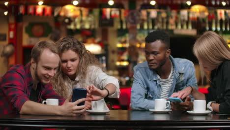 Multiethnische-Paare-Sitzen-In-Einem-Café,-Schauen-Auf-Die-Bildschirme-Ihrer-Telefone-Und-Blättern-Fotos-Auf-Dem-Telefonband-Durch.