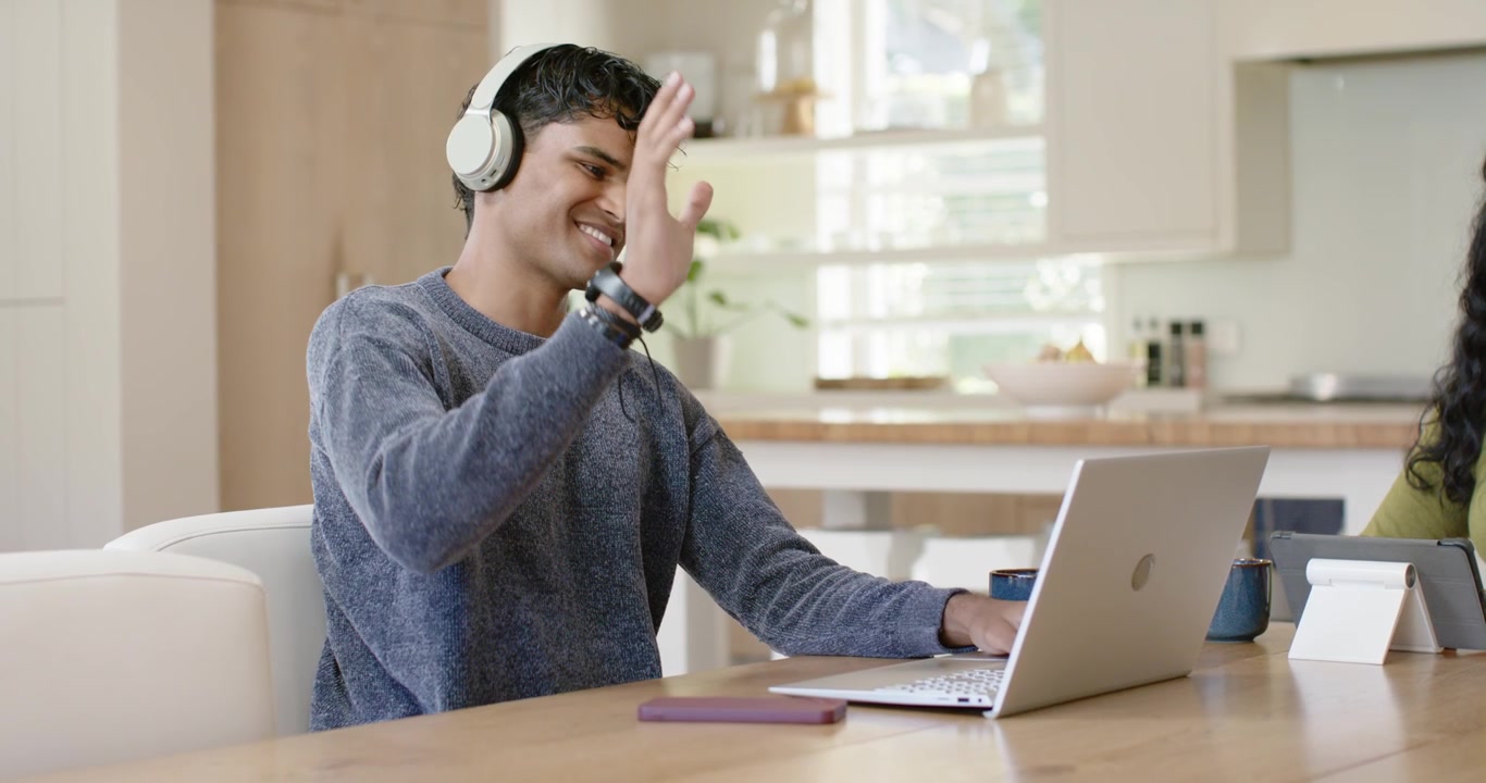 Mangfoldigt par i køkkenet, mand iført hvidt headset skriver på bærbar computer, tager opkald og vinker