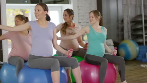 group of four fit pregnant women training with dumbbells while sitting on stability balls in fitness class