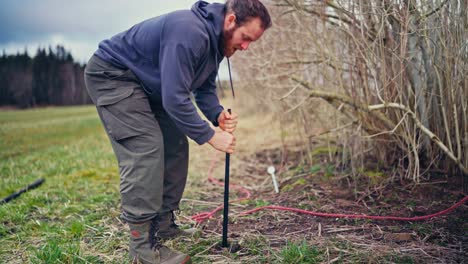man drills hole for wooden post while constructing fence - wide shot