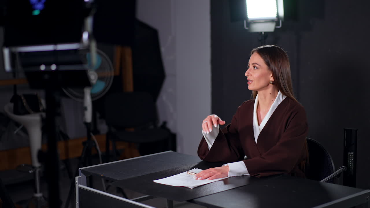 Attractive Caucasian woman with long brown hair sits at desk in studio. Positive lady talks smiling.