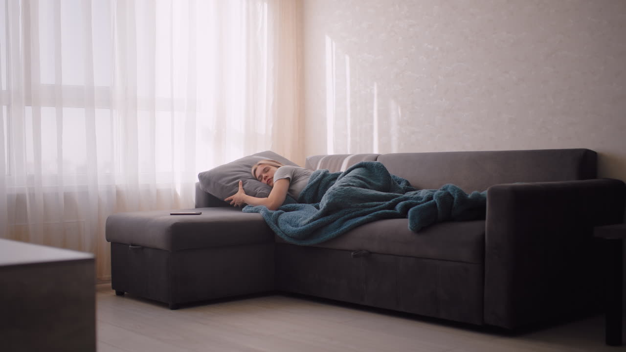 Young woman resting on couch holding pillow wrapped in blanket with phone nearby as soft sunlight enters through sheer curtains peaceful scene showing calm atmosphere quiet home environment