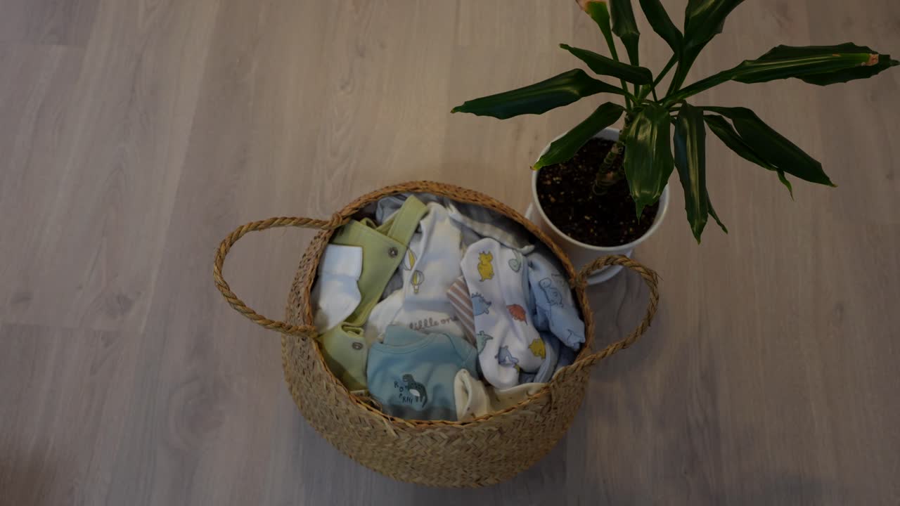 Wicker basket filled with folded baby clothes placed on wooden floor, overhead view