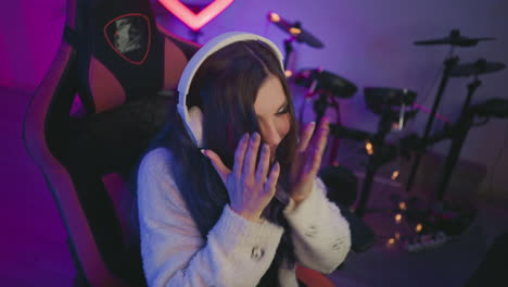 elegant woman with long wavy hair wearing white headphones leans forward smiling while engaging with unseen task, seated in gaming chair with drum set and colorful lights in studio background