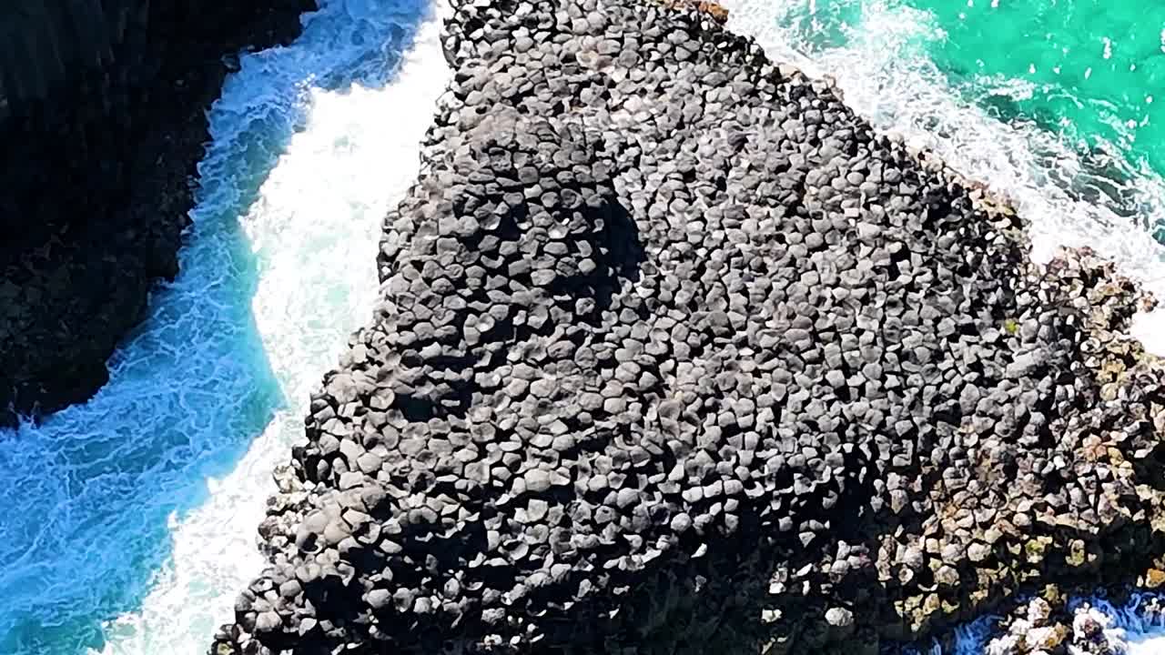 Close-up aerial view of hexagonal basalt columns meeting turquoise ocean waves.