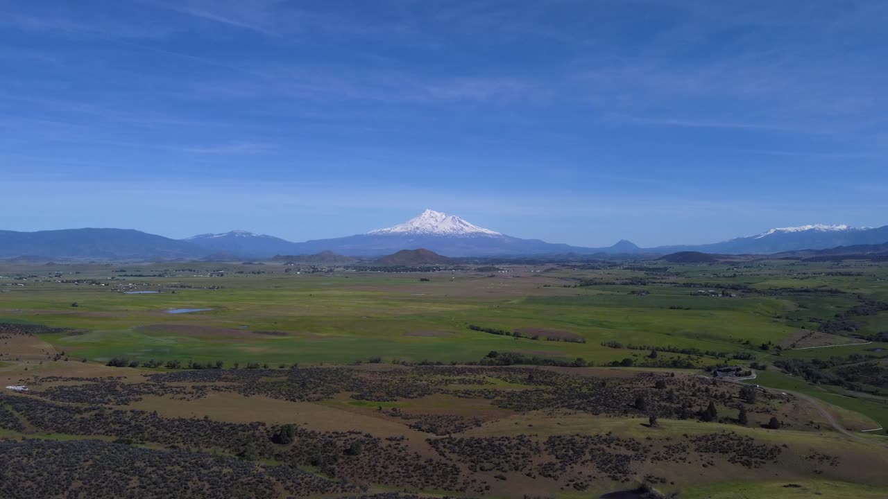 Mount Shasta from a green, grassy, agricultural valley in California. The mountain towers over the valley and provides a stunning contrast of the white, glacier laden peak and blue sky. 4K drone shot.