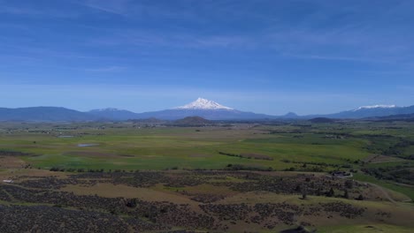 mount shasta from a green, grassy, agricultural valley in california. the mountain towers over the valley and provides a stunning contrast of the white, glacier laden peak and blue sky. 4k drone shot.