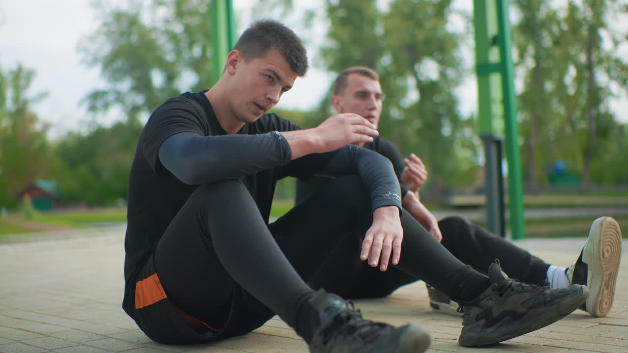 Caucasian athletes sitting on pavement resting, two young caucasian men under park shelter after intense run, catching breath, checking watch, rubbing temples, sharing quiet conversation, green trees
