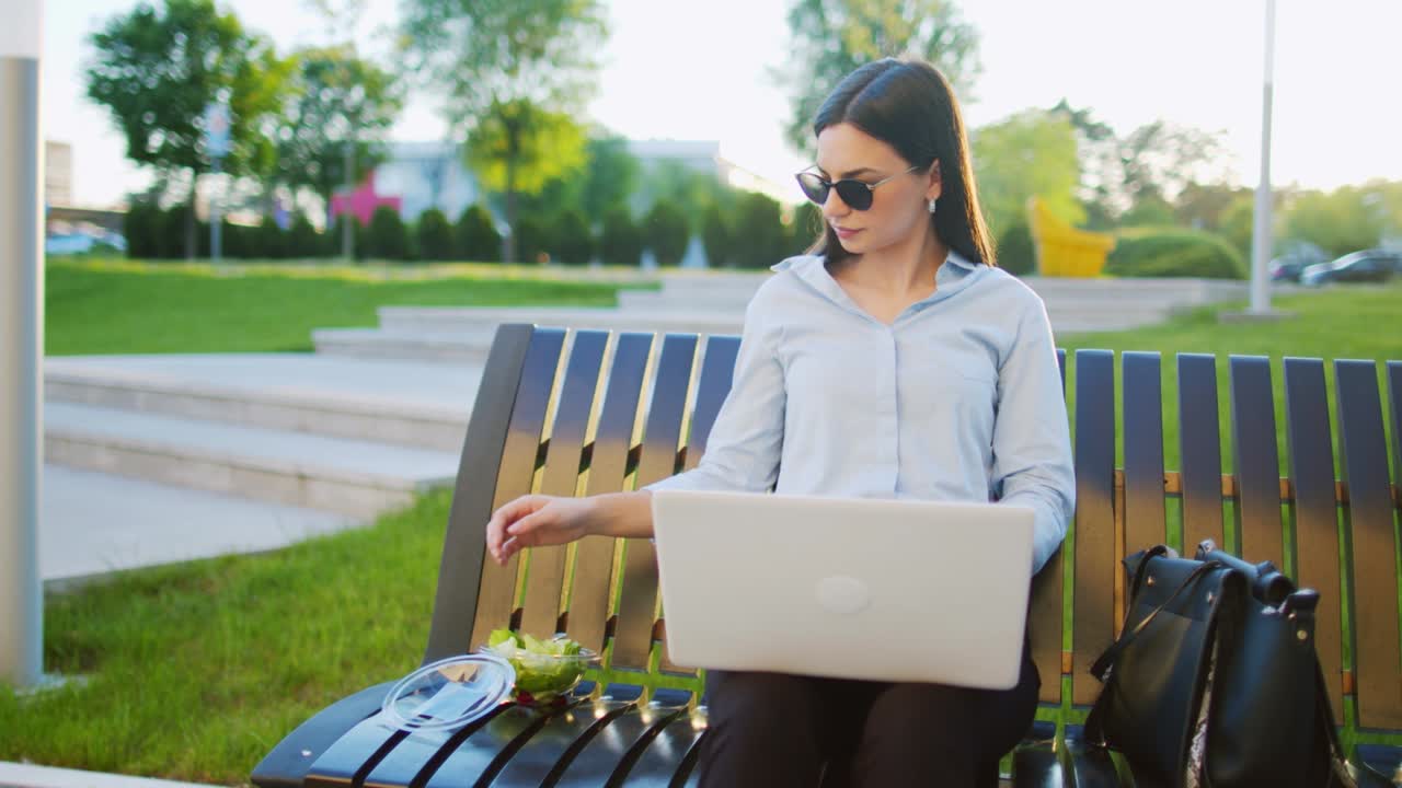 Mujer de negocios sentada en un banco al aire libre usando una computadora portátil para trabajar fuera de la oficina y comiendo ensalada.