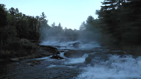 Aerial-drone-shot-over-a-dark-misty-forest-stream-and-waterfall-at-sunset-with-trees-in-silhouette