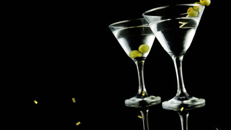 golden confetti falling over olives in two cocktail glasses against black background