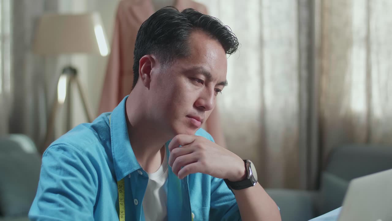 스튜디오에서 옷을 디자인하는 동안 노트북에서 일하고 생각하는 아시아 남성 디자이너의 클로즈업