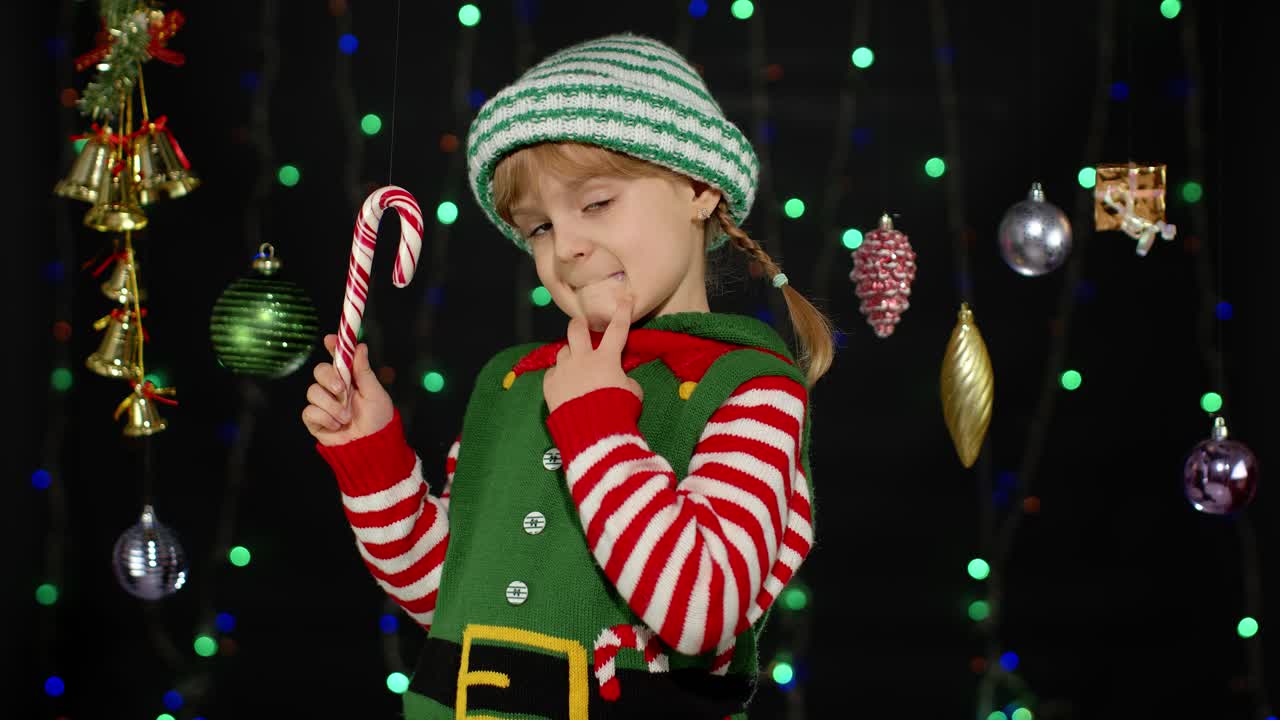 Kid girl in Christmas elf Santa Claus helper costume with candy cane lollipop joyful smiling