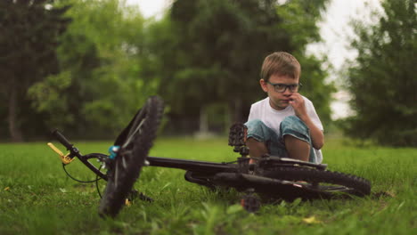 a young boy squats next to his bicycle, removed something on his face in a grassy field with blurred greenery in the background
