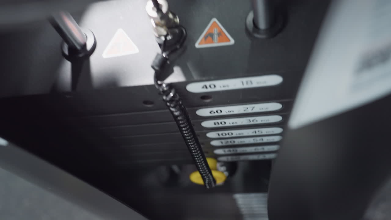 Close up of weight stack moving on gym machine showing pin shifting between plates labeled in pounds and kilograms under bright gym lighting with cable coil and selector pin adjusting resistance