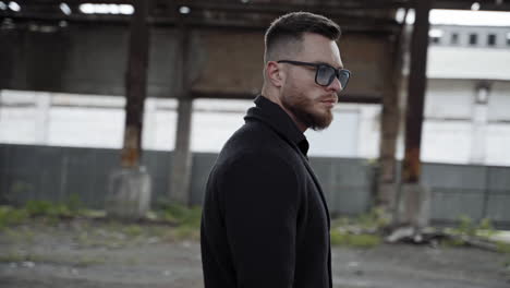 handsome man in ruined place. serious bearded man in glasses stands with baseball bat inside the damaged building. camera moves in around.