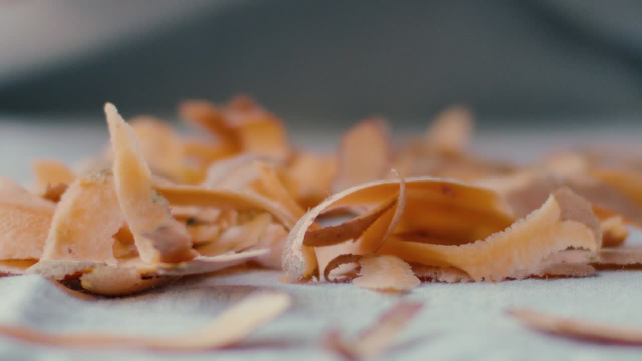 A sweet potato is peeled with a potato peeler in the background, in the foreground detail of the peels piling up on the table falling in slow motion