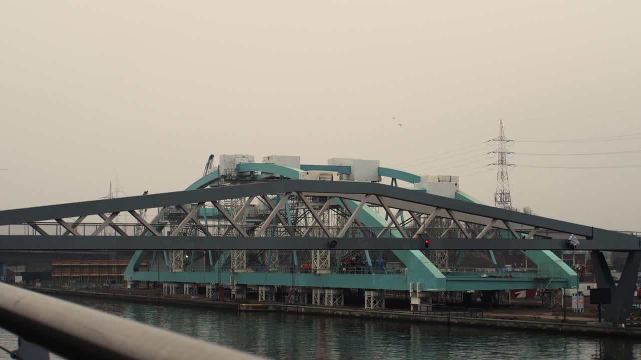 Dual bridge scene with a small pedestrian and bicycle bridge in the foreground and a large aqua-colored bridge under construction in the background, showcasing layers of urban infrastructure