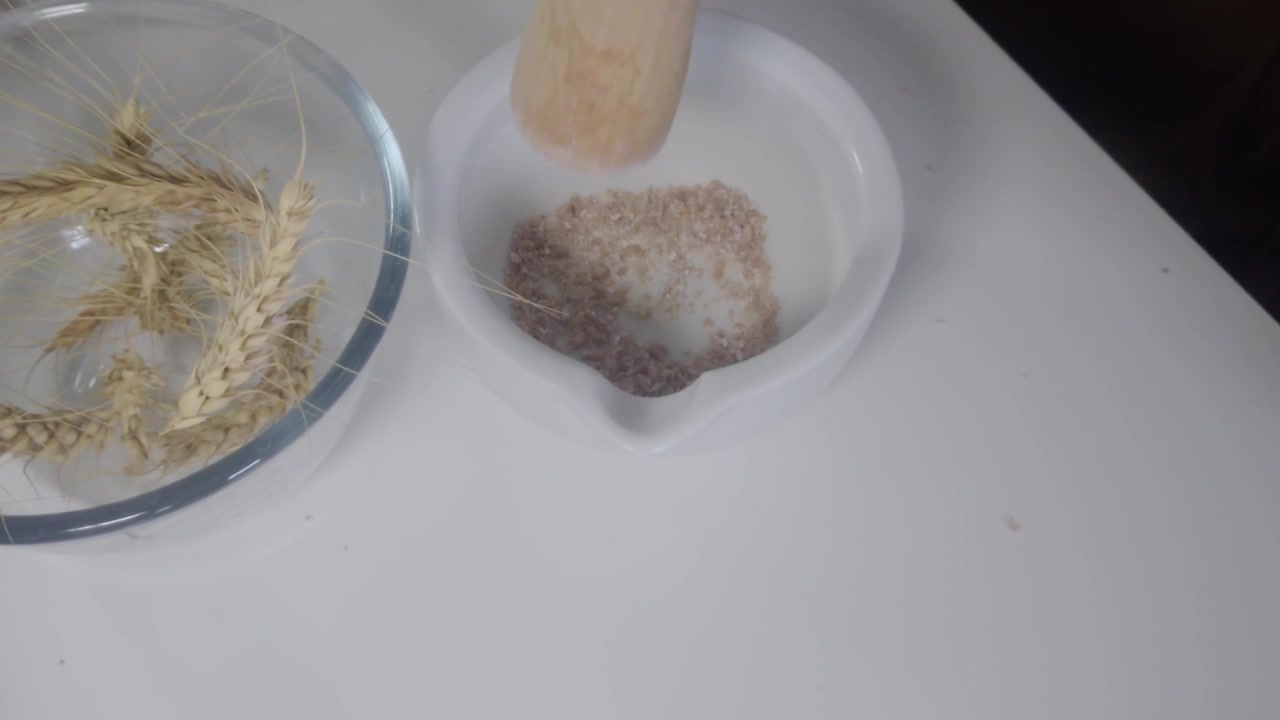 Un'immagine sfocata dall'alto, morbida, mostra mani sfocate che macinano chicchi di grano con un pestello di legno in un mortaio accanto a una ciotola di vetro, creando una texture di movimento astratta ideale come sfondo per video educativi