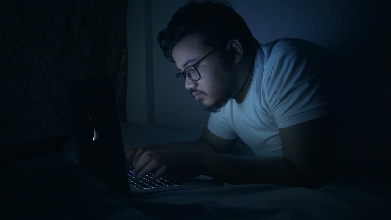 인디언 남자가 침대에 노트북을 사용하여 오버타임을 하고 있습니다.