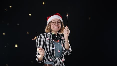 una linda mujer con gorro de papá noel y coloridas guirnaldas y auriculares en el cuello con dos bengalas bailan en la fiesta de la noche de navidad sobre fondo negro. mujer joven sonriente y alegre divirtiéndose
