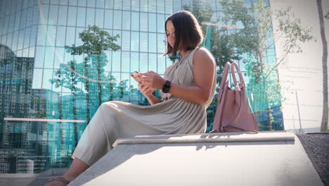 Business-woman-working-in-the-courtyard-surrounded-by-high-rise-office-buildings-with-glass-facade-on-her-smartphone