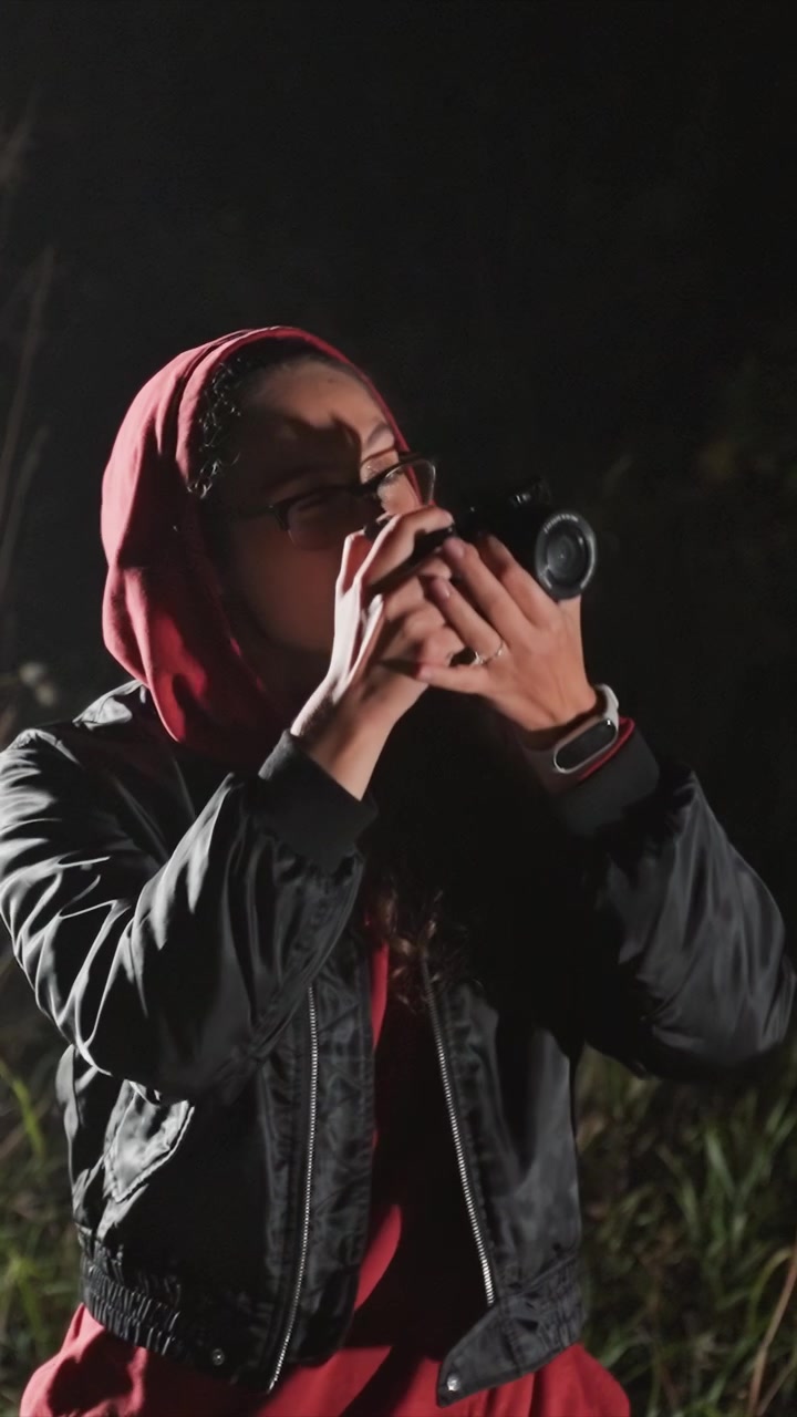 Young woman in red hoodie and black jacket holds camera. Woman keeps binoculars in place on left wrist with watch. Outdoor area shows grass behind subject under low light
