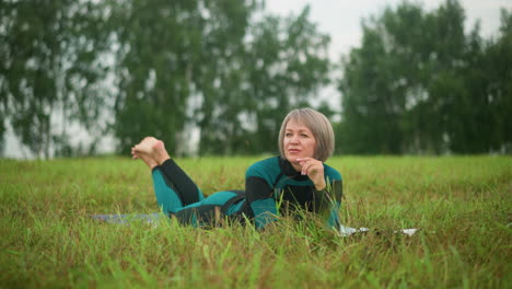 woman lying on grassy field with legs slightly raised and hand resting under chin, gazing into distance with a warm smile, surrounded by trees in background