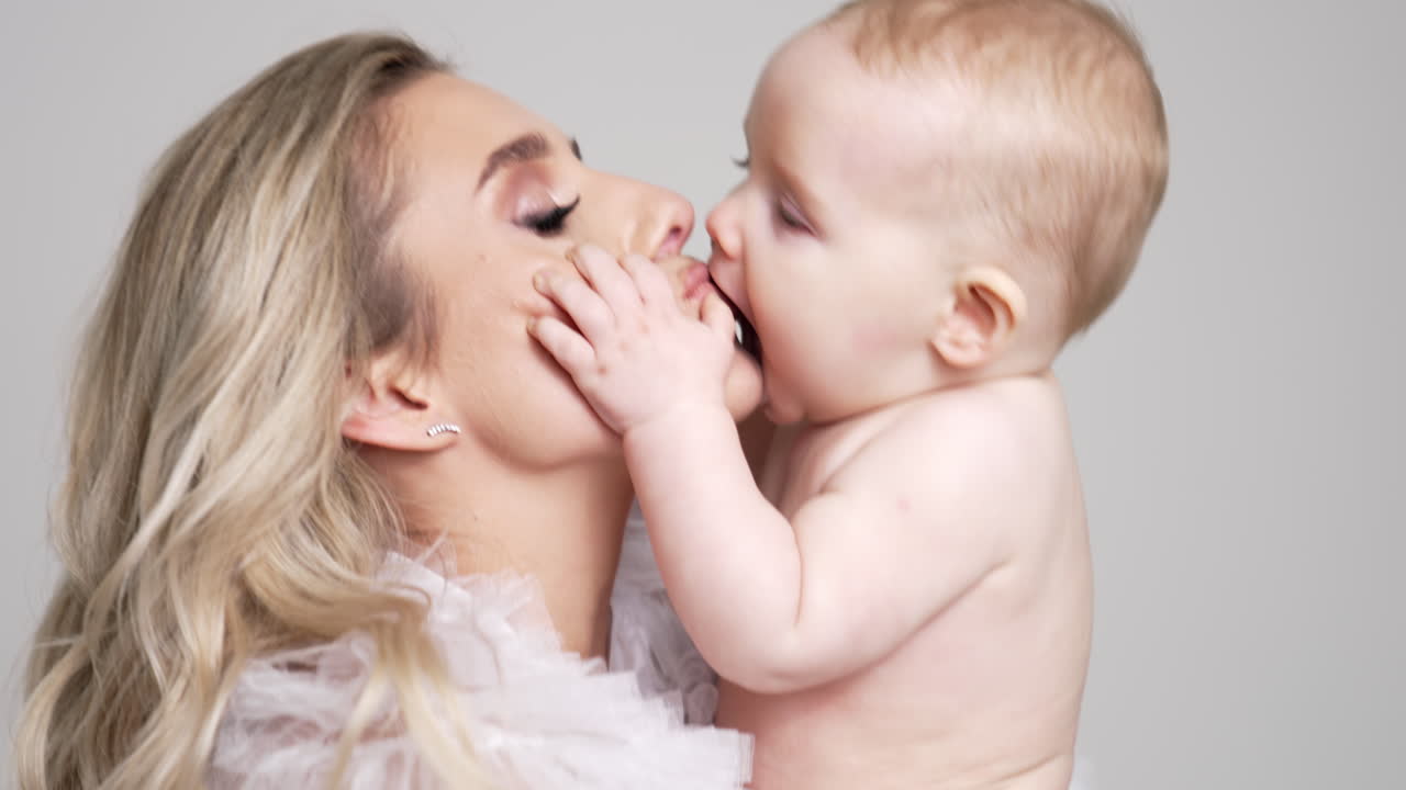 Caucasian blonde woman holds her naked infant baby. Mom jumps up with her son and kisses him with love. White backdrop.