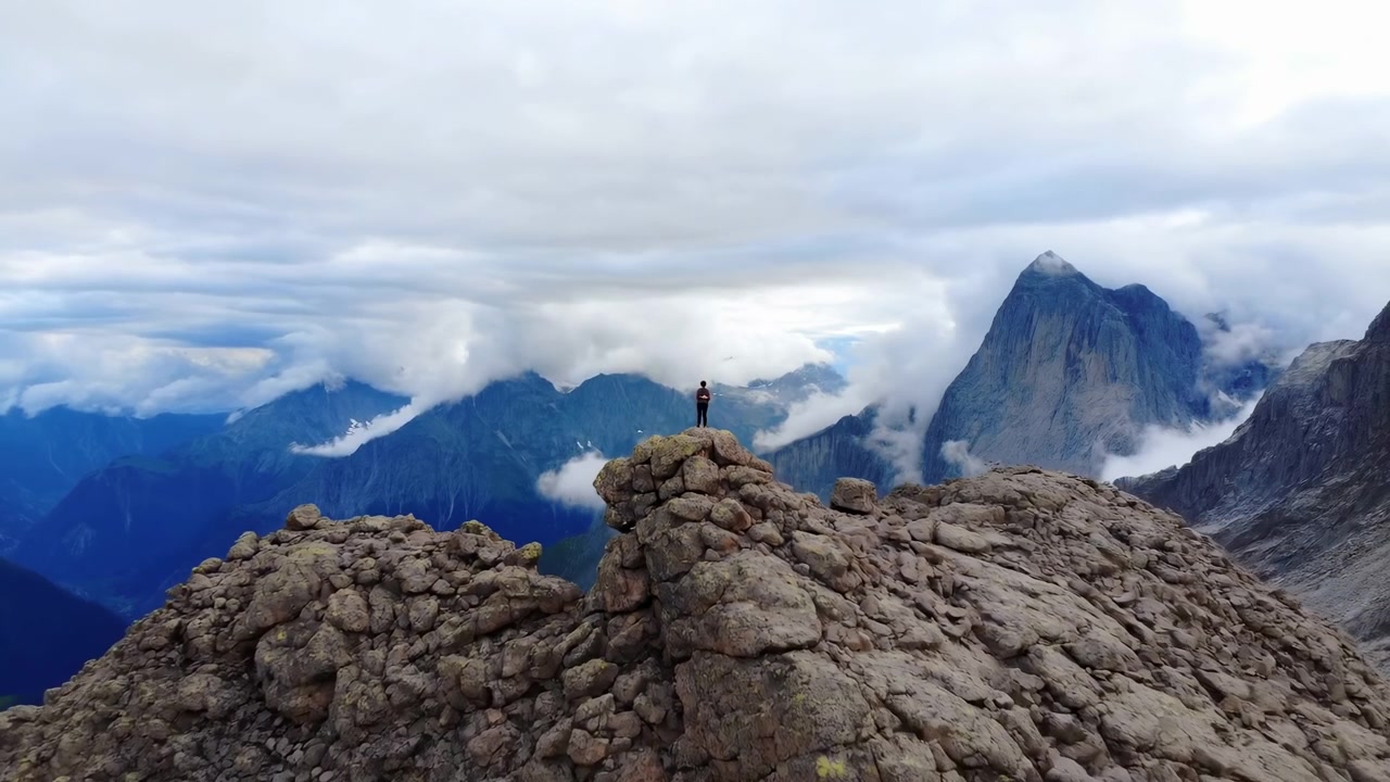 Escursionista solitario in cima a una vetta rocciosa, vista panoramica sulle montagne, strati di nuvole drammatici che scorrono attraverso una valle alpina