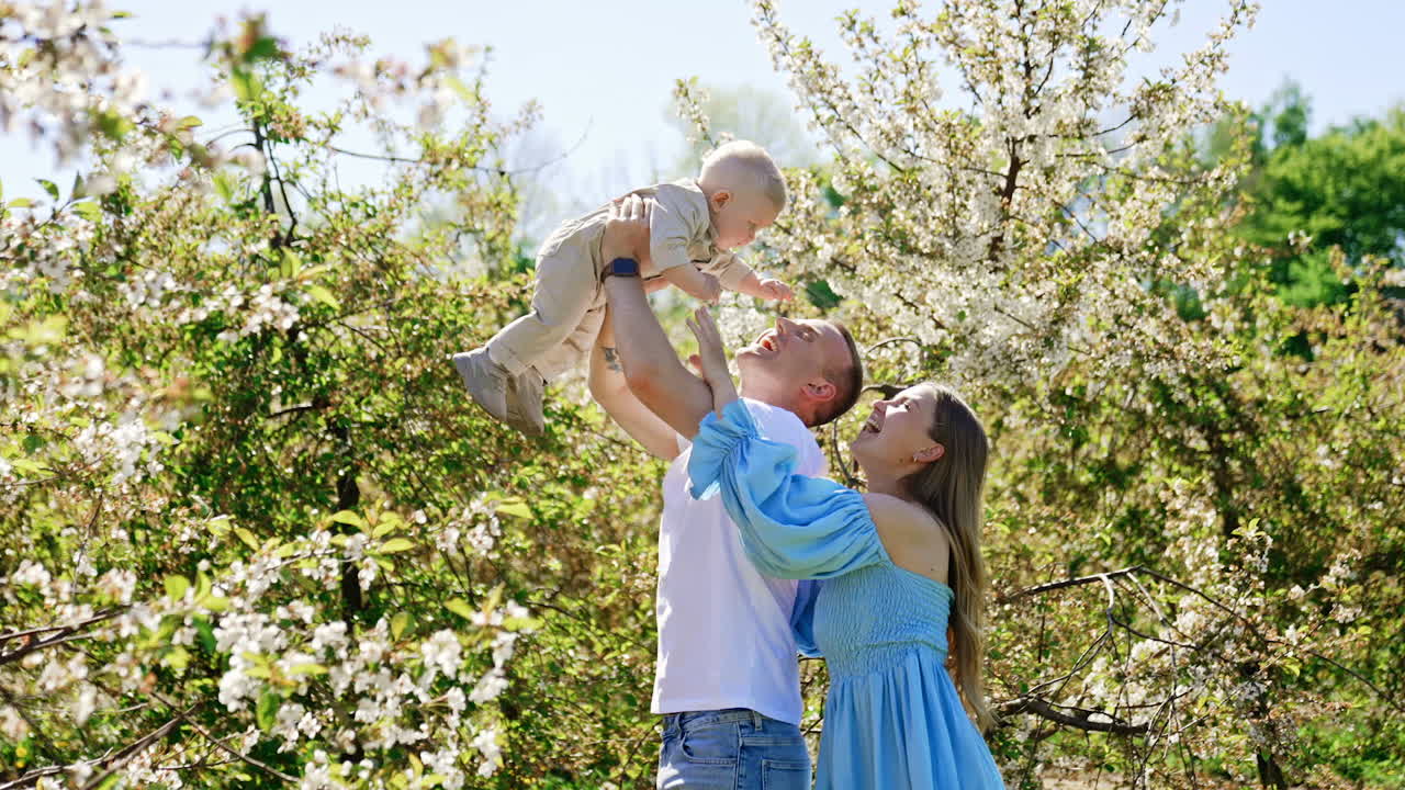 Caucasian man holding his little baby boy high above the head. Mom stands behind her husband smiling to the kid. Spring garden at backdrop.