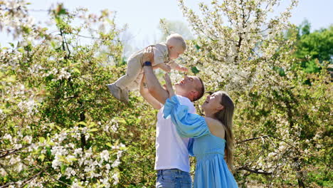 caucasian man holding his little baby boy high above the head. mom stands behind her husband smiling to the kid. spring garden at backdrop.
