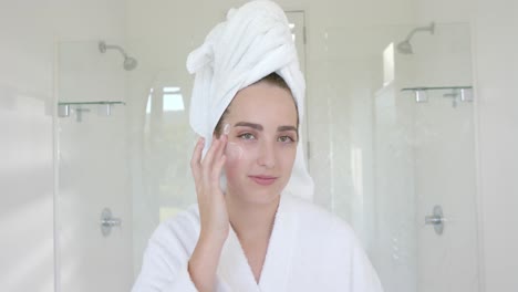 Portrait-of-caucasian-woman-with-towel-on-head-applying-cream-on-face-in-bathroom-in-slow-motion