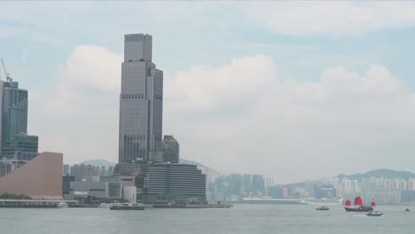 a view of victoria harbour with a red-sailed junk boat inspired by ancient chinese ships, now a tourist attraction, sailing alongside other boats against the backdrop of the kowloon modern skyline.