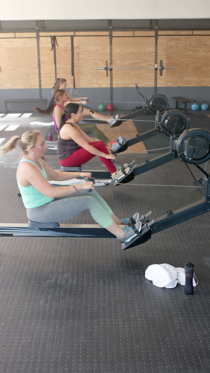 Vertical video of diverse sportswomen exercising with rowing machines at gym in slow motion