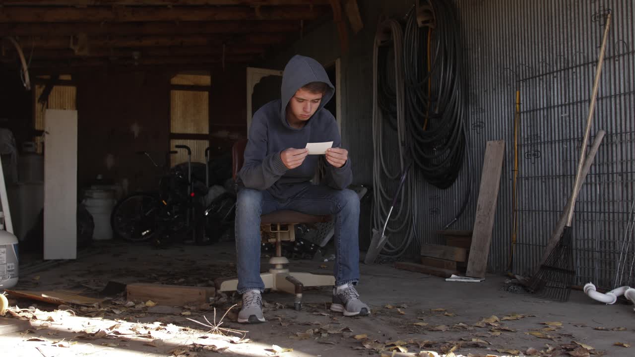 A teenage boy sits in a dirty garage looking at a picture