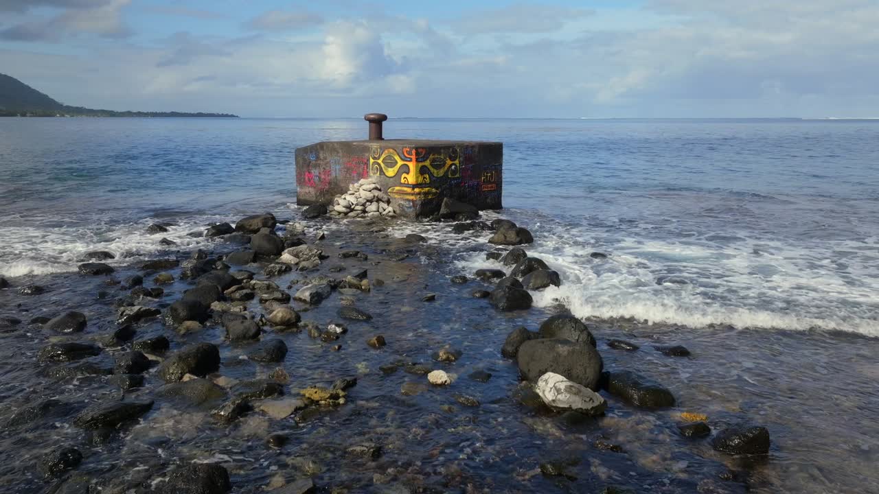 Street art by the sea Taiarapu road to Teahupoo Tahiti French Polynesia drone South Pacific Ocean cement block Polynesian face graffiti painting low tide rocks waves crashing sunny upward jib shot