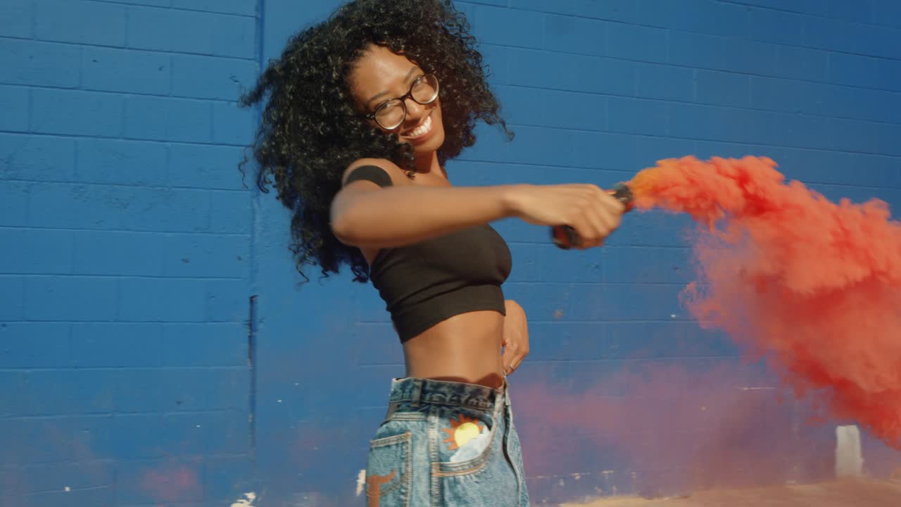 Beautiful young woman holding colorful smoke grenade dancing outside against blue wall