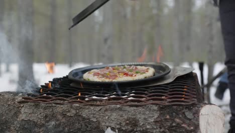 removing a steel lid to show a pizza being cooked on a steel grate over an open wood fire