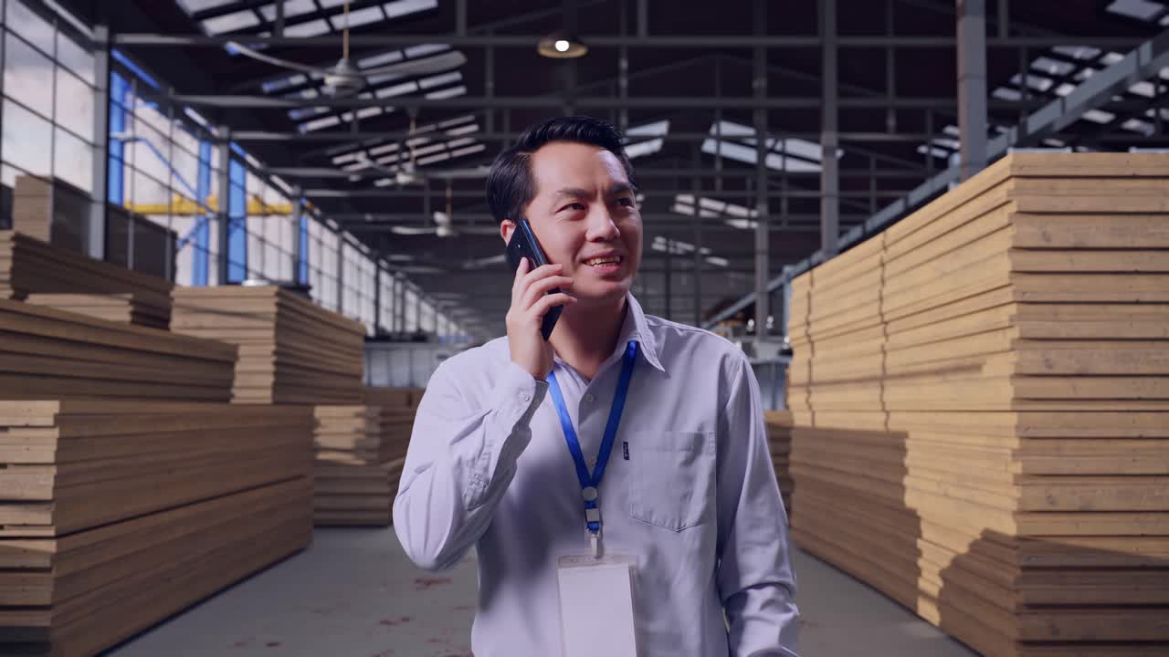 Operaio asiatico professionista in piedi che ispeziona un magazzino di legname, prende il telefono e inizia a parlare.
