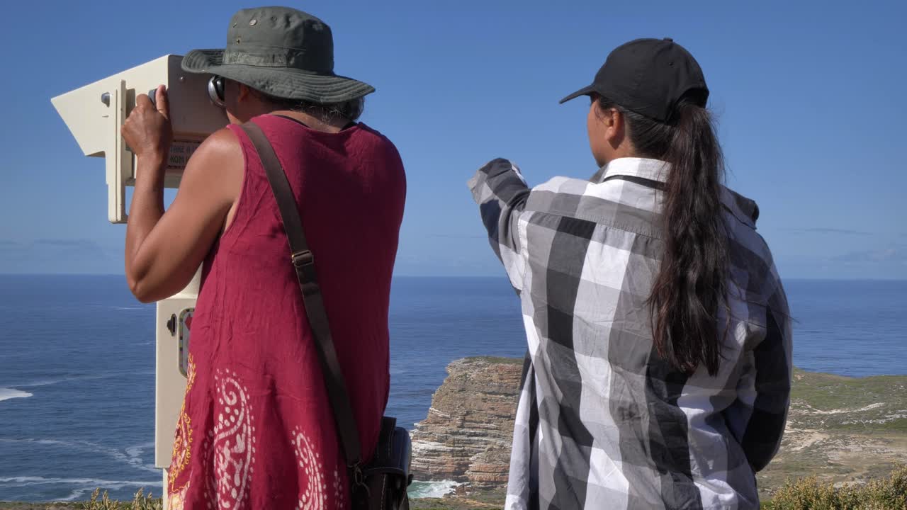 Due donne, una che indica l'oceano, usano binocoli a gettoni per ammirare il vasto oceano e il paesaggio costiero frastagliato a Cape Point, Penisola del Capo, Sudafrica