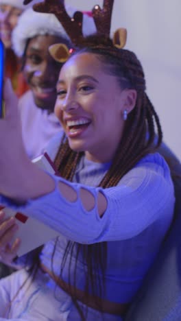 Vertical-Video-Studio-Shot-Of-Gen-Z-Friends-At-Christmas-Sitting-On-Sofa-Wearing-Santa-Hat-And-Reindeer-Antlers-Taking-Selfie-On-Mobile-Phone-1