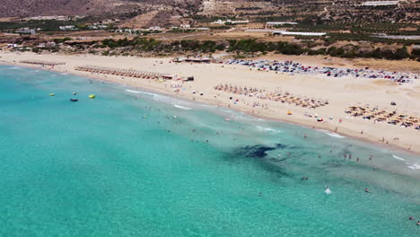 aerial view descending to falassarna beach with tourist sunbathing on sandy greek island and swimming in transparent clear ocean water