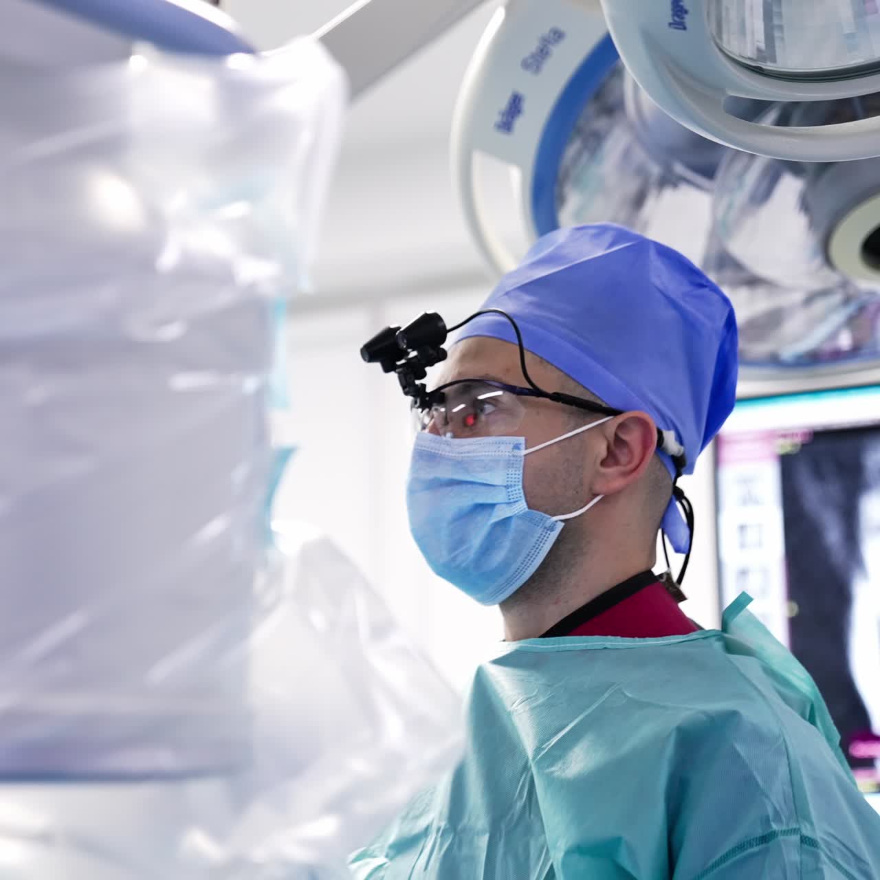 Man surgeon with special device on his head stands in the operation room. Medical professional holds instrument in his hand and looks in front of him